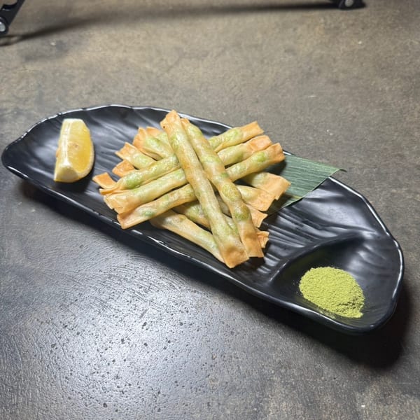 Crispy fried spring rolls with green filling, a lemon wedge, and green powder on a black plate.