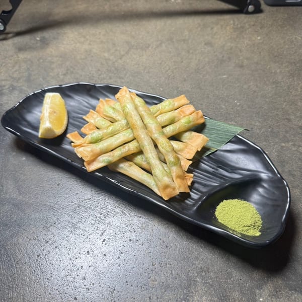 Crispy green bean tempura sticks with a lemon wedge and green matcha powder on a black plate.