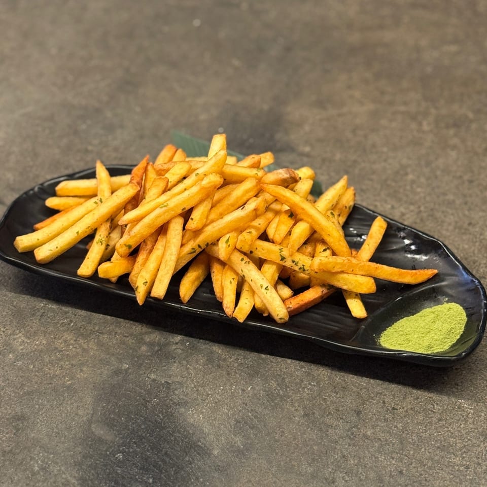 Golden french fries seasoned with herbs, served on a black plate with a side of green powder.