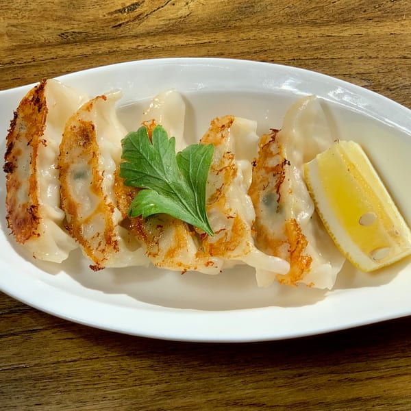 A white plate of pan-fried HINODEYA gyoza with a lemon wedge and parsley on a wooden table.